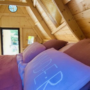 Cabane en bois en Bretagne, lit douillet avec coussins colorés et vue sur la nature.