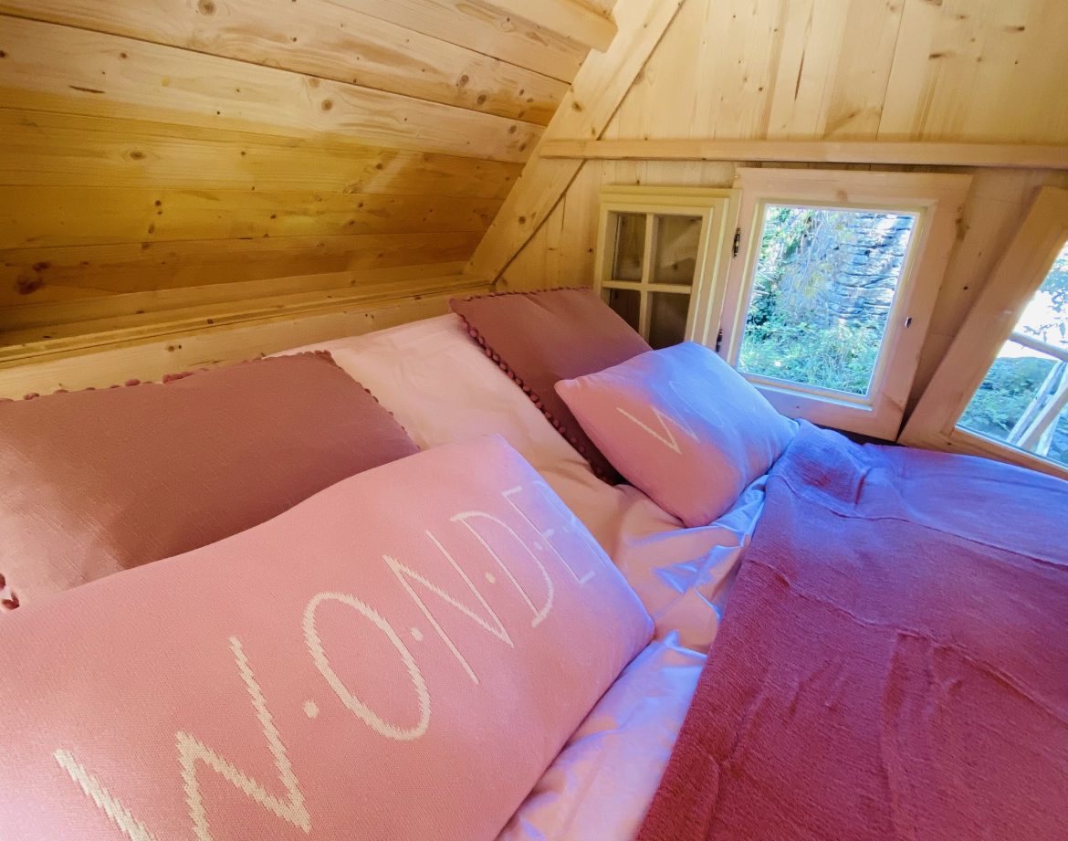 Cabane en bois en Bretagne, lit douillet avec coussins roses et vue sur la nature.