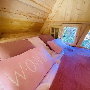 Cabane en bois en Bretagne, lit douillet avec coussins roses et vue sur la nature.