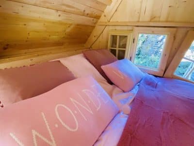 Cabane en bois en Bretagne, lit douillet avec coussins roses et vue sur la nature.