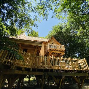 Cabane en bois perchée, entourée darbres, avec une grande terrasse en bois.