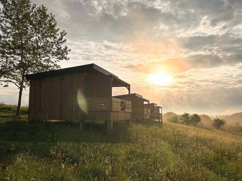 Cabines en bois au coucher de soleil, entourées de verdure et dun ciel nuageux.