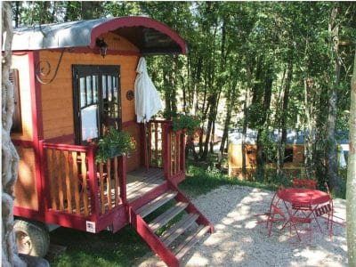 Roulotte colorée en bois, entourée darbres, avec terrasse et mobilier extérieur.