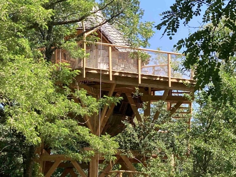 Cabane perchée dans les arbres, entourée de verdure luxuriante et dun ciel bleu.