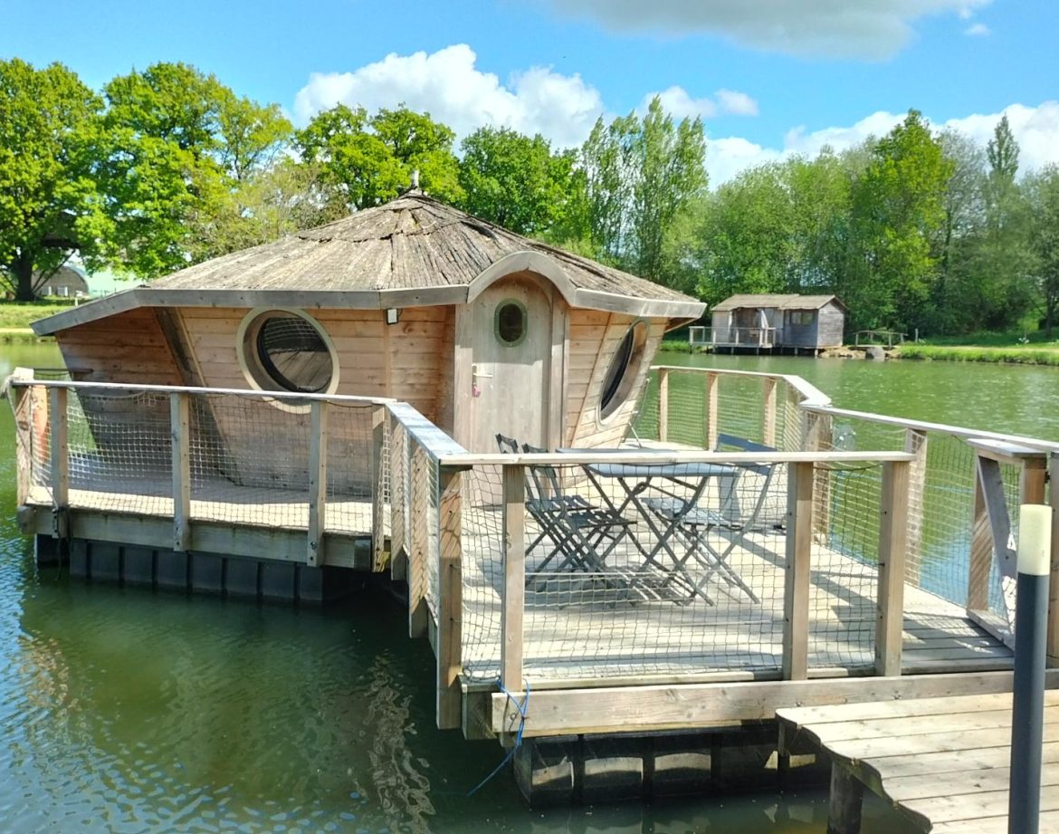Cabane flottante en bois avec terrasse sur un étang, entourée de verdure.