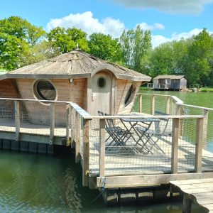 Cabane flottante en bois avec terrasse sur un étang, entourée de verdure.