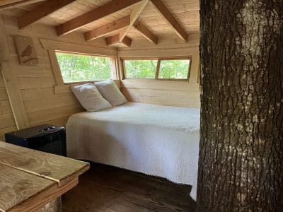 Cabane perchée dans les arbres, intérieur en bois avec lit douillet et grandes fenêtres.