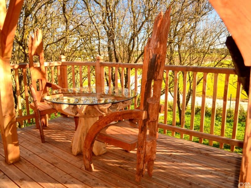 Cabane perchée avec terrasse en bois et table en rondins, vue sur la nature.