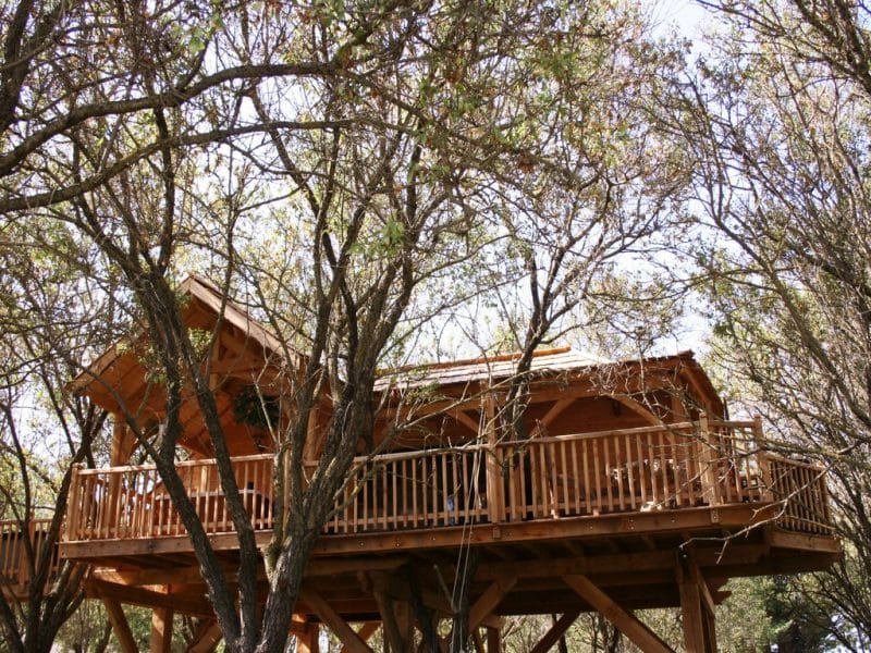 Cabane dans les arbres en bois, entourée de feuillage verdoyant et dune terrasse accueillante.