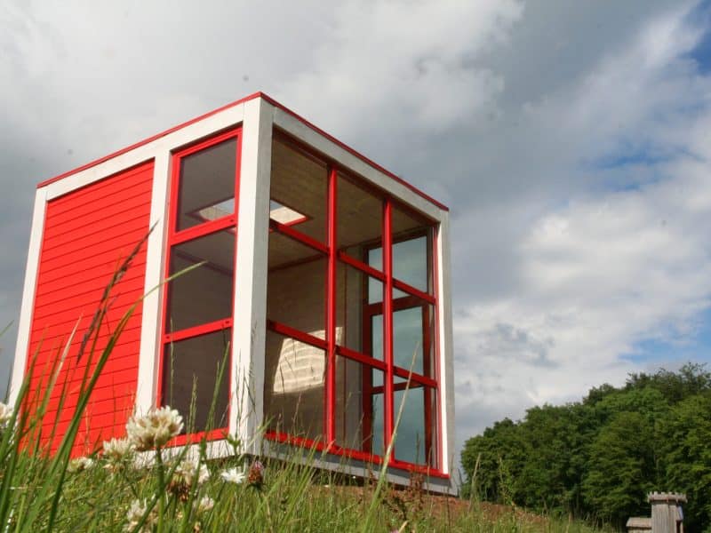 Cabane moderne en bois rouge, avec de grandes fenêtres et vue sur la nature environnante.