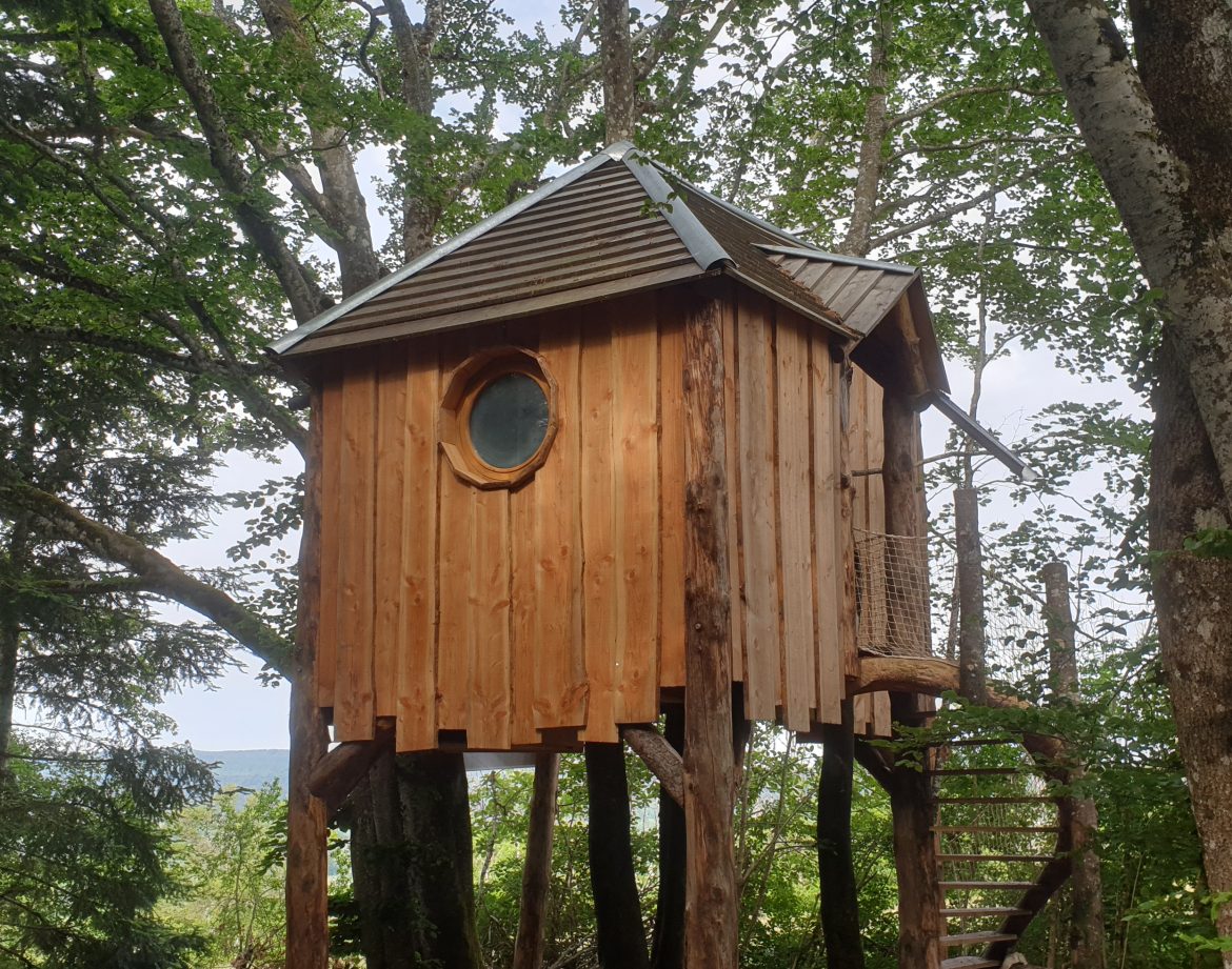 Cabane perchée en bois dans les arbres, avec une fenêtre ronde, en Auvergne-Rhône-Alpes.