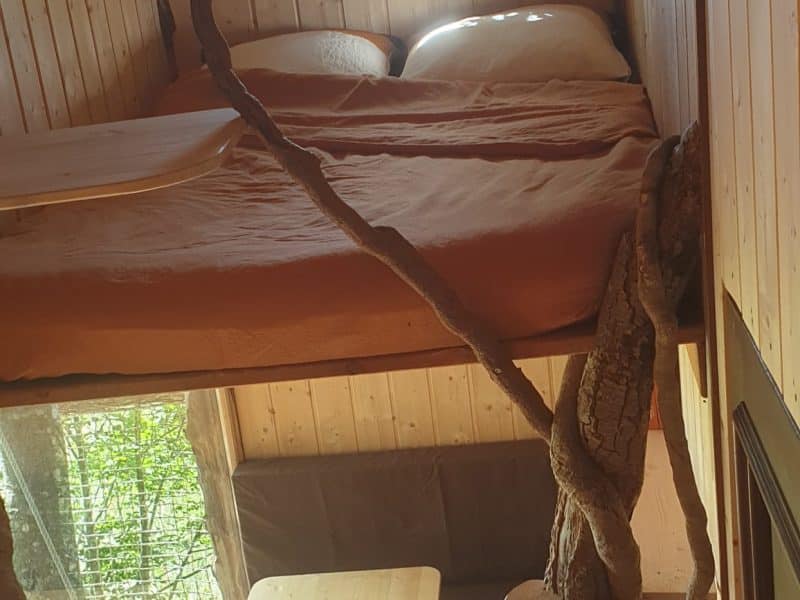 Cabane perchée en bois avec lit douillet et escalier en branches naturelles.