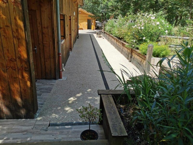Chalet en bois avec allée en gravier, entouré de verdure et de fleurs.