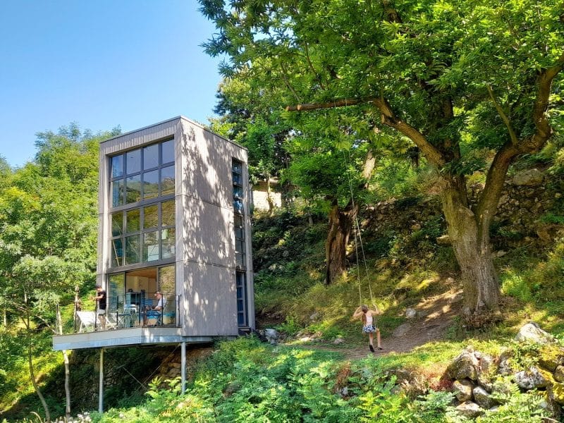 Cabane moderne en bois, entourée de verdure, avec grandes fenêtres et terrasse.