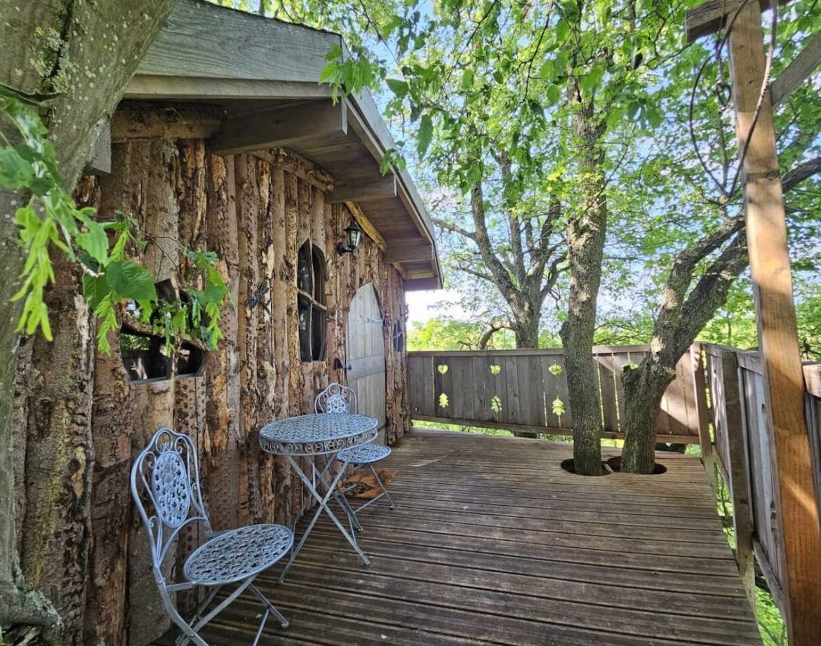 Cabane perchée en bois, avec terrasse en bois et mobilier en fer forgé sous les arbres.