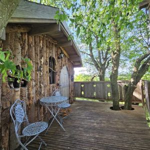 Cabane perchée en bois, avec terrasse en bois et mobilier en fer forgé sous les arbres.
