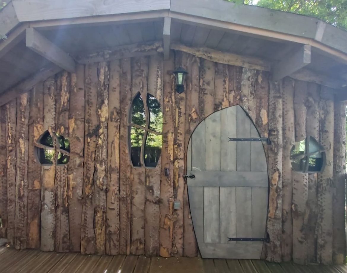 Cabane en bois dans les arbres, avec des fenêtres en forme darc et une porte en bois.