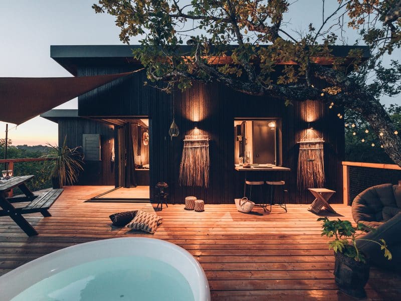 Cabane moderne en bois avec terrasse, baignoire extérieure et éclairage chaleureux.