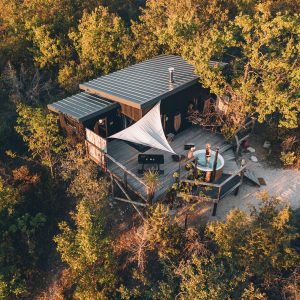 Cabane perchée en bois entourée de verdure, avec une terrasse et un coin détente.