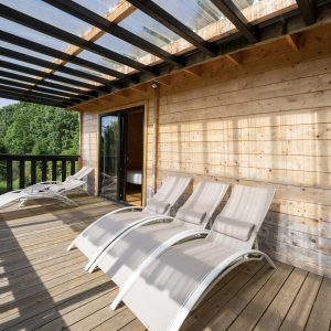 Cabane en bois avec terrasse ensoleillée et chaises longues, vue sur la nature.