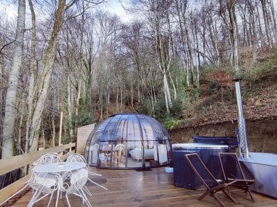 Dôme transparent en bois, entouré darbres, avec jacuzzi et chaises blanches.