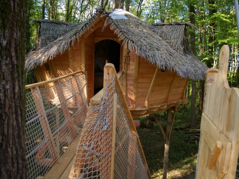 Cabane perchée en bois avec toit de chaume, accessible par un pont suspendu.