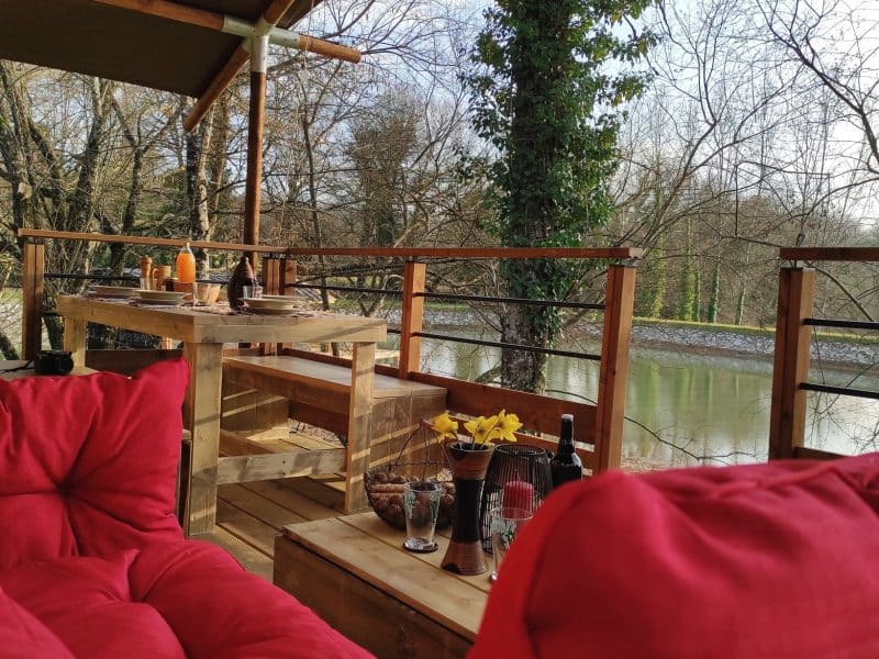 Hébergement en cabane avec terrasse, vue sur la rivière et coussins rouges confortables.