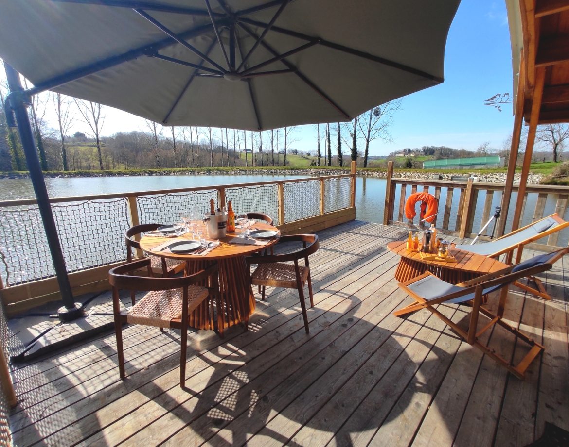 Cabane sur pilotis au bord de leau, avec terrasse en bois et parasol.
