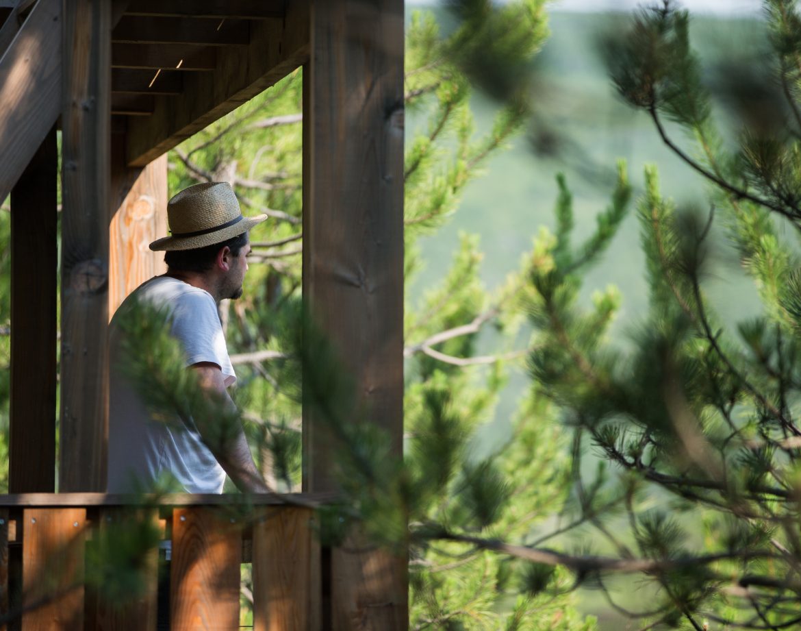 Cabane perchée en bois avec vue sur la nature, homme en chapeau sur le balcon.
