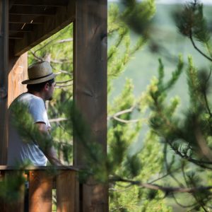 Cabane perchée en bois avec vue sur la nature, homme en chapeau sur le balcon.