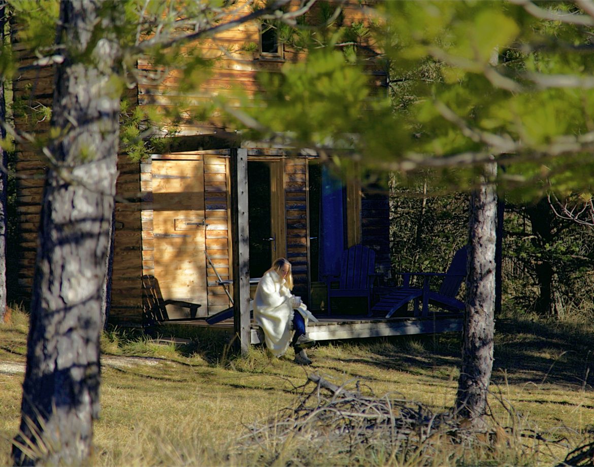 Cabane en bois dans la nature, avec une personne assise sur la terrasse.