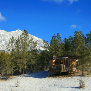 Cabane perchée en bois, entourée de sapins et face à des montagnes enneigées.