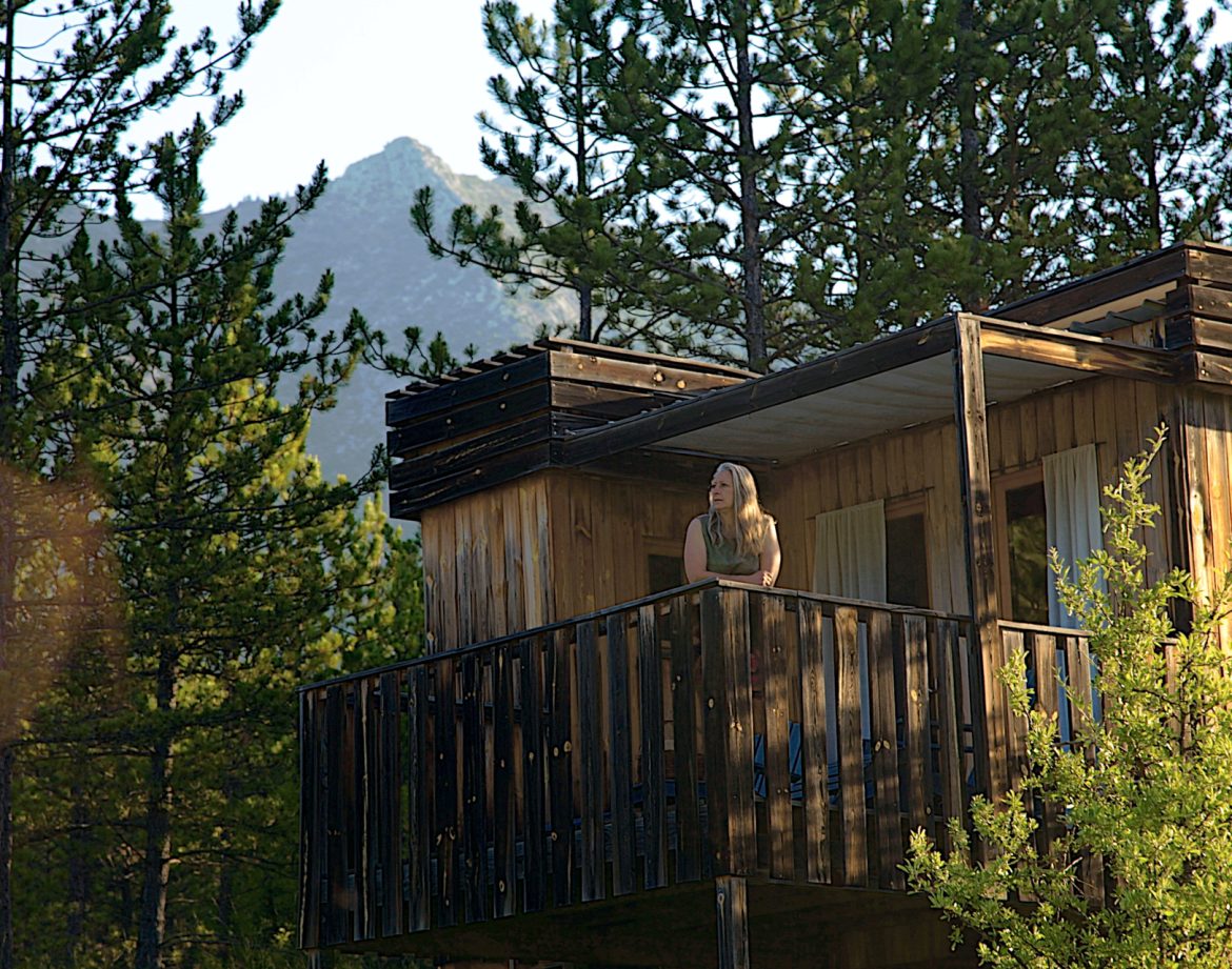 Cabane perchée en bois avec balcon, entourée de pins et vue sur les montagnes.