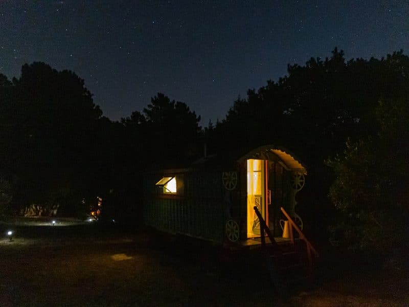 Charmante roulotte illuminée sous un ciel étoilé, entourée de verdure.