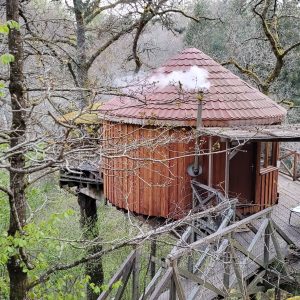 Cabane perchée en bois dans les arbres, avec un toit en tuiles et cheminée fumante.