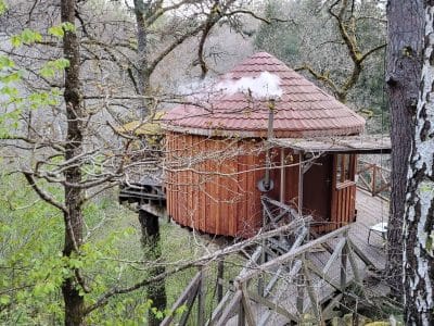 Cabane perchée en bois dans les arbres, avec un toit en tuiles et cheminée fumante.