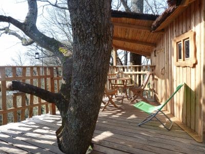 Cabane perchée dans un arbre avec terrasse en bois et vue sur la nature environnante.