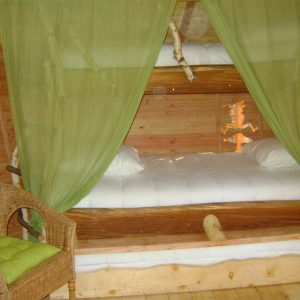 Cabane perchée avec lits en bois, drapée de rideaux verts et fauteuil en osier.