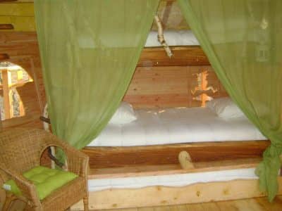Cabane perchée avec lits en bois, drapée de rideaux verts et fauteuil en osier.