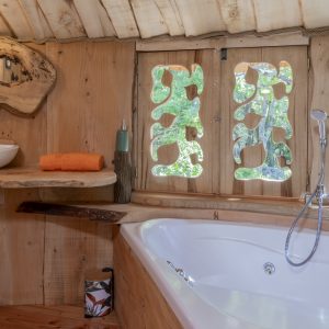 Cabane en bois avec baignoire, fenêtres sculptées et vue sur la nature.