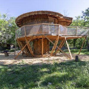 Cabane perchée en bois, entourée de verdure, avec une terrasse en bois sur pilotis.