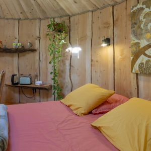 Hébergement insolite en cabane boisée, lit douillet avec coussins colorés et décoration naturelle.