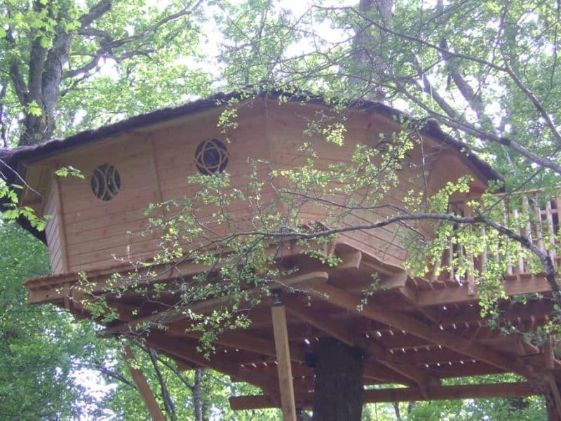 Cabane perchée dans les arbres, avec des fenêtres rondes et un accès en bois.
