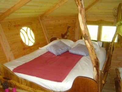 Cabane en bois avec lit douillet et fenêtres arrondies, ambiance chaleureuse.