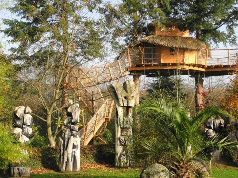 Cabane dans les arbres avec toit de chaume, entourée de sculptures en bois.