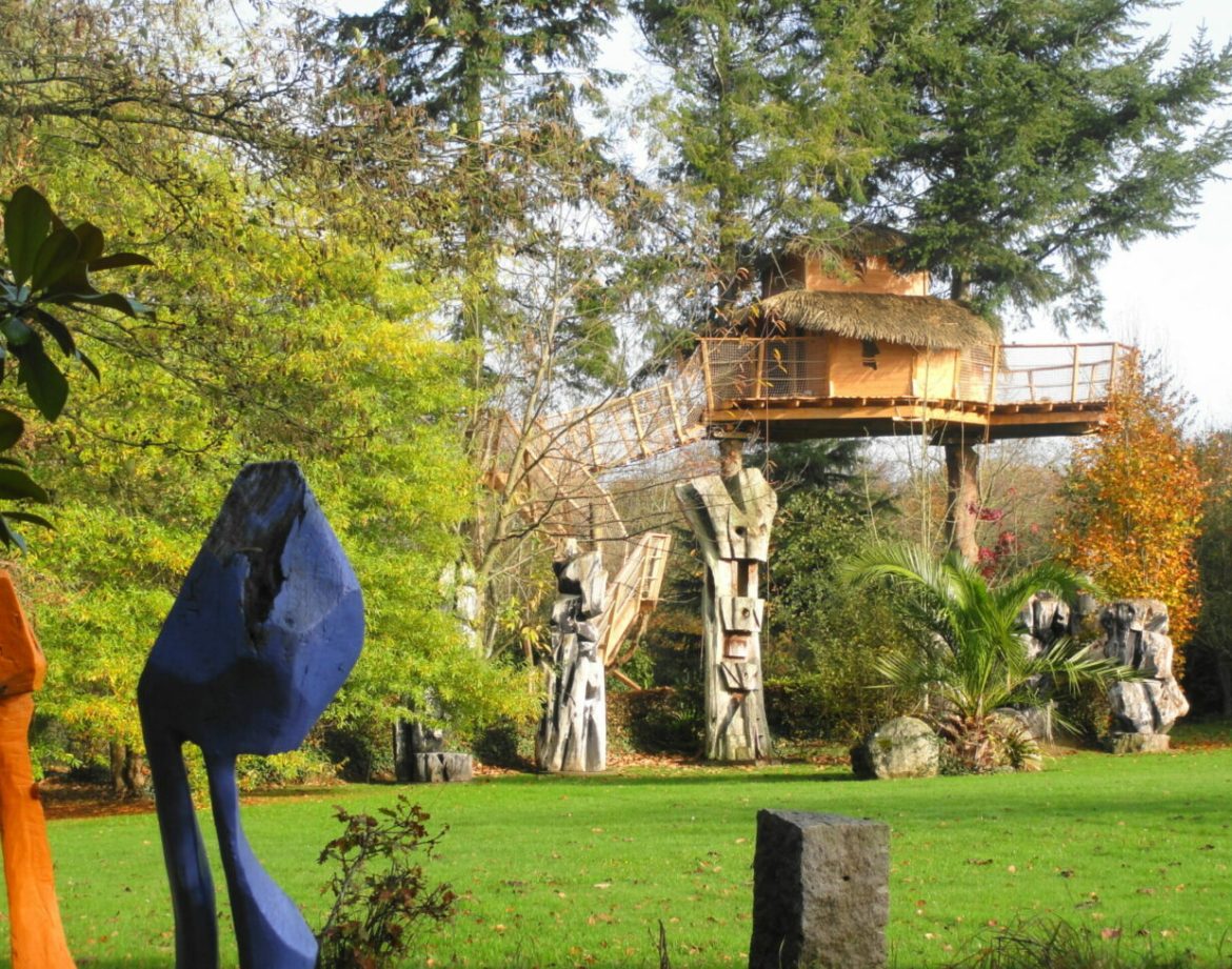 Cabane perchée en Bretagne, entourée de sculptures colorées et de verdure luxuriante.