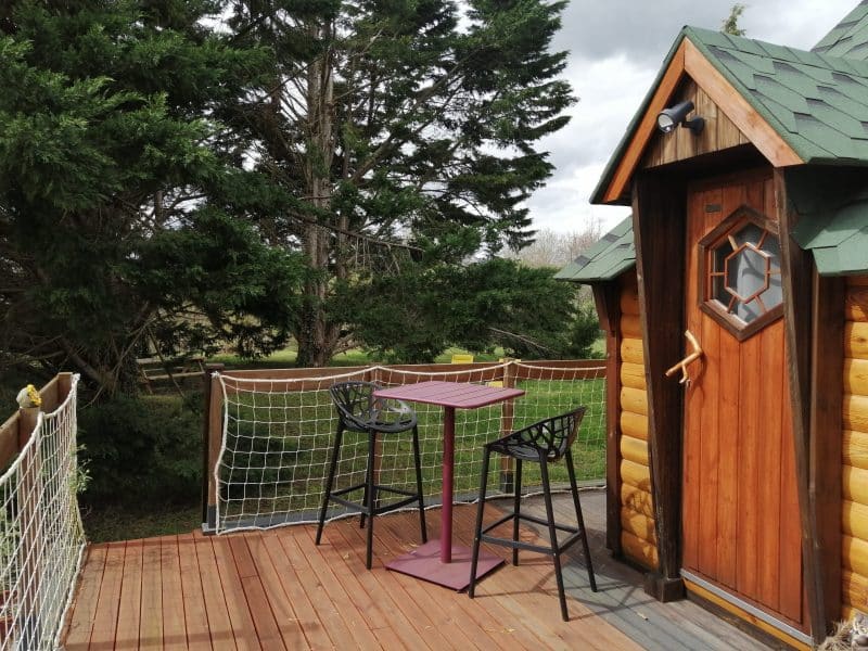 Cabane en bois avec terrasse et chaises, entourée de verdure.
