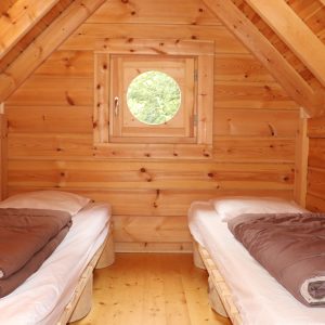Chalet en bois avec deux lits simples et une fenêtre ronde, ambiance chaleureuse.