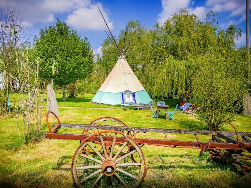 Tipi coloré en pleine nature, entouré darbres et dun ancien chariot en bois.