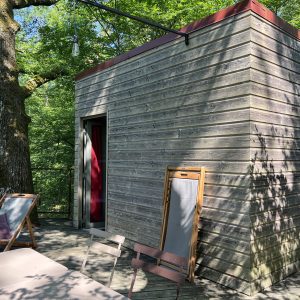 Cabane en bois perchée, entourée darbres, avec terrasse en bois et mobilier extérieur.